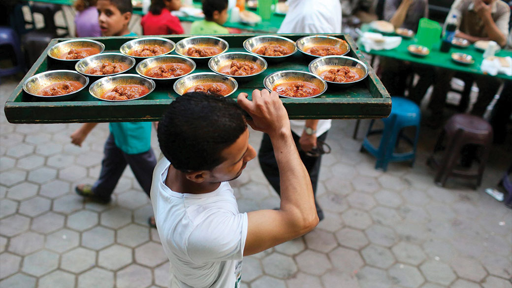 A volunteer carries food as people wait to eat their iftar meal to break their fast at charity tables that offer free food during the holy fasting month of Ramadan in Cairo, Photo courtesy of Reuters.